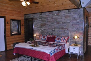 A bedroom with a stone wall, wooden ceiling, a bed with a quilt, two bedside tables with lamps, and a door. - Secluded, luxurious cabin perched above Eagle Fork Creek with sunrise views. (Smithville)