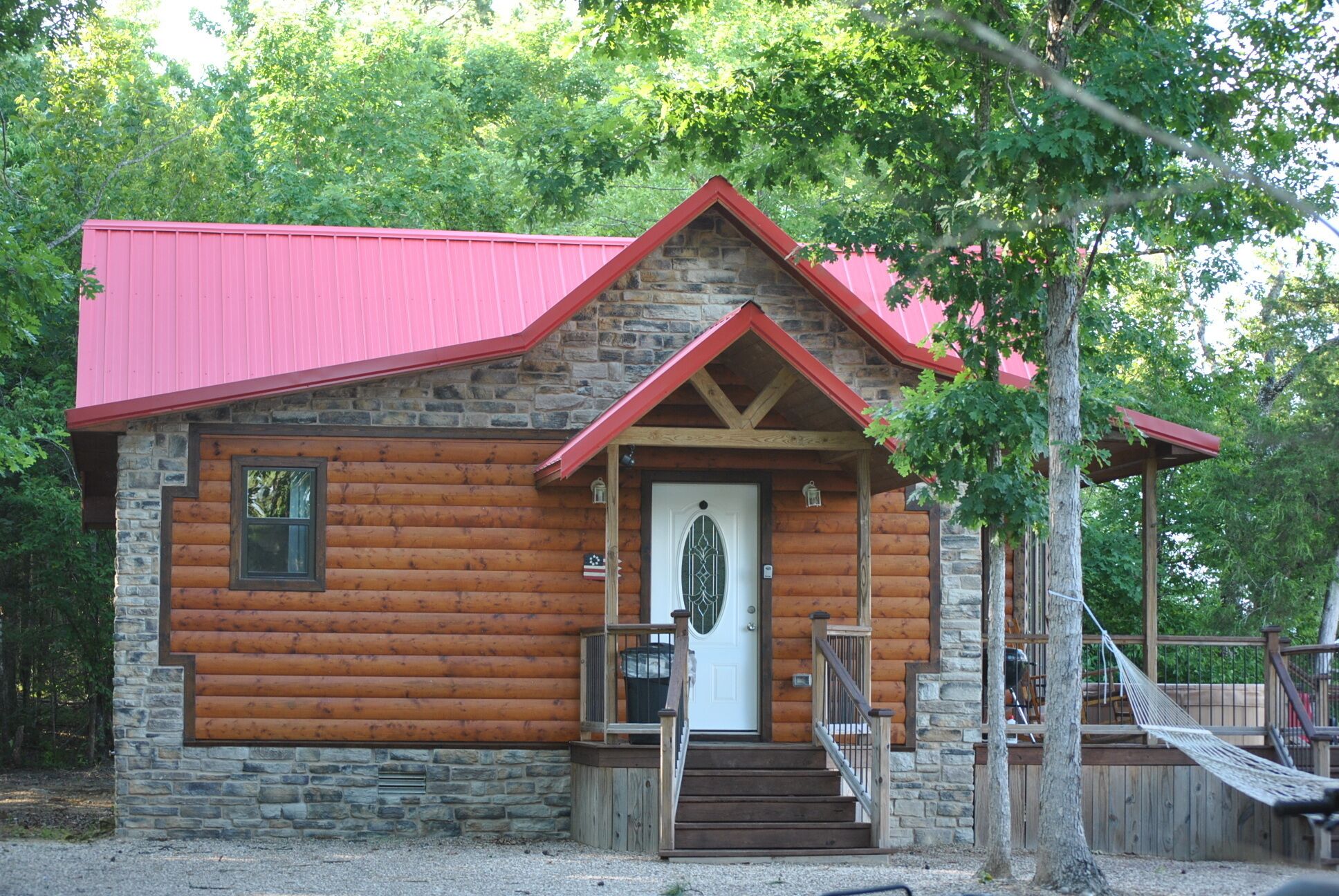 A log cabin with a stone foundation, a red roof, and a front porch with steps leading to a white door.