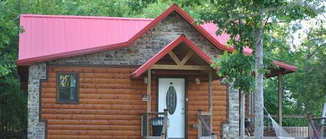 A log cabin with a stone foundation, a red roof, and a front porch with steps leading to a white door.