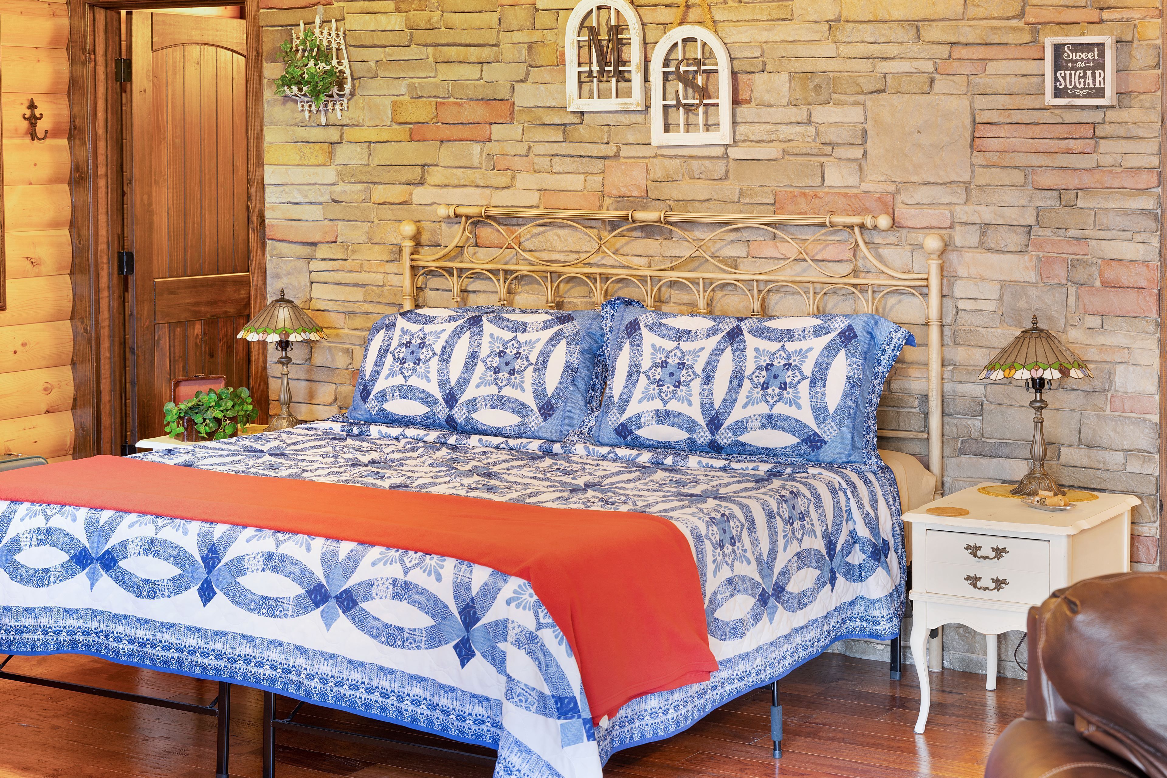 A bedroom with a wooden door, a stone wall, a bed with blue and white bedding, a nightstand, and a lamp.