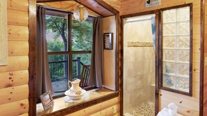 A wooden cabin interior with a large window, a wooden bench, a small table with a pitcher and a picture frame, and a towel on a wooden stool.
