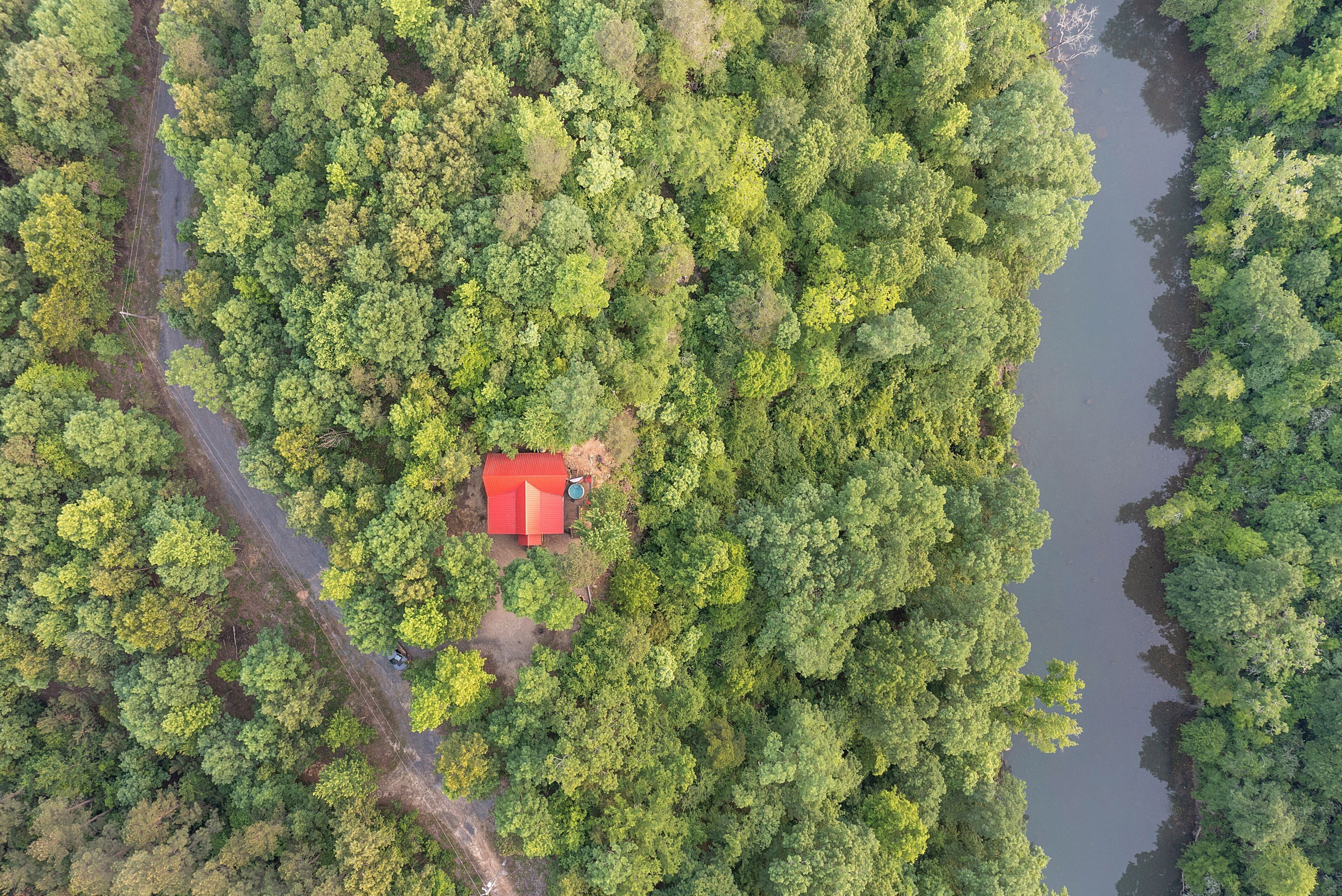 An aerial view of a forested area with a small red-roofed building surrounded by dense trees and a water body.
