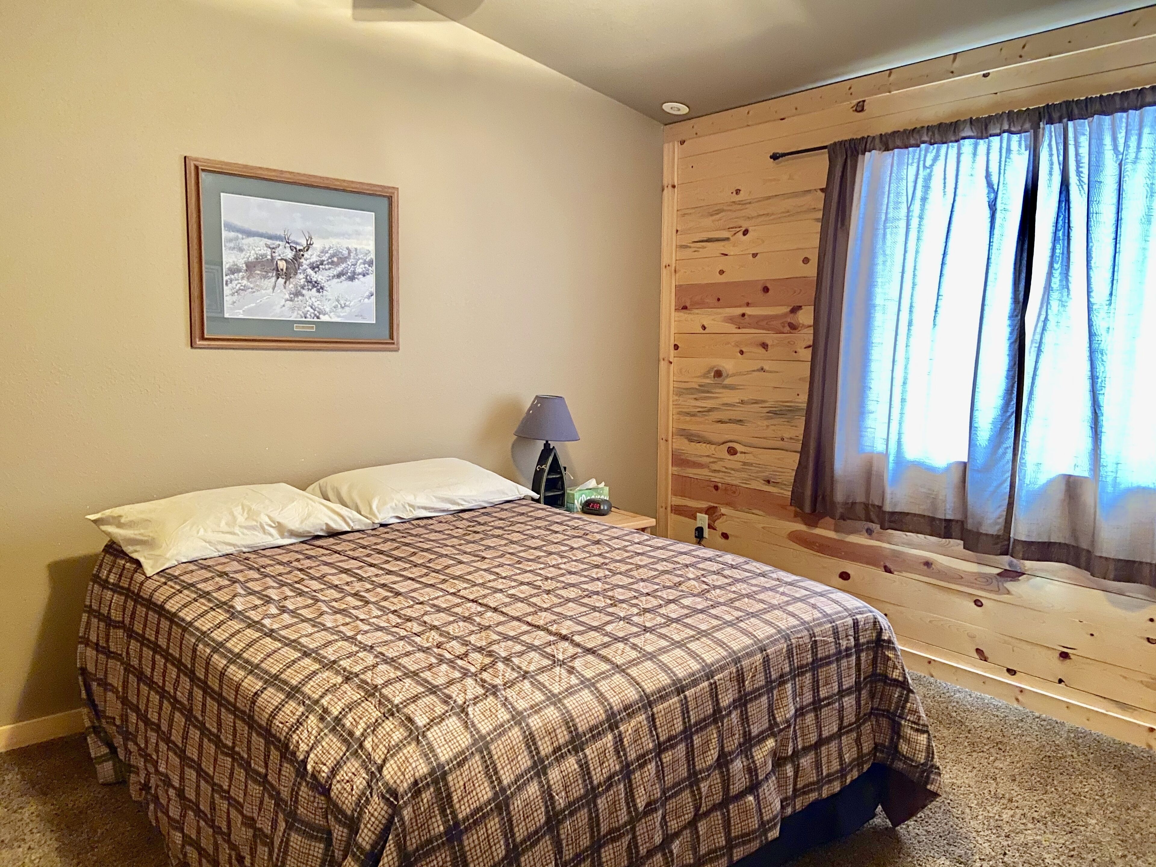 A bedroom with a plaid bedspread, a framed picture on the wall, a lamp, and a window with curtains.