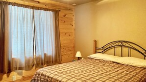 Une chambre avec un plafond en bois, un lit avec une tête de lit en métal, une table de chevet avec une lampe et une fenêtre avec des rideaux.