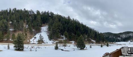 A snowy landscape with a forested mountain in the background and a wooden deck with chairs.