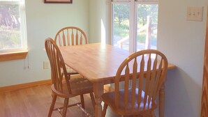A dining area with a wooden table and four chairs, a framed artwork on the wall, and a window with a view of greenery. - Mimi's Mansion At Stacy Ranch, Cabin On 120 Acres Between Ingram & Hunt, Tx (Ingram)