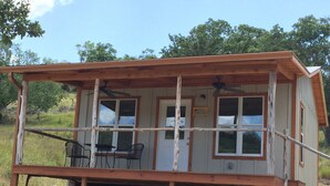 A rustic cabin with a wooden deck, two windows, and a small table with chairs. - Mimi's Mansion At Stacy Ranch, Cabin On 120 Acres Between Ingram & Hunt, Tx (Ingram)
