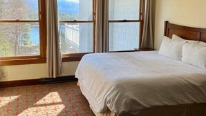 Ein Hotelzimmer mit einem großen Bett, zwei Fenstern mit Vorhängen, einer Deckenleuchte und Blick auf ein Gebäude und Bäume.