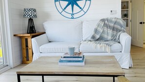 Un salon bien éclairé, avec un canapé blanc, une table basse sur laquelle sont posés des livres, une boussole fixée au mur et une petite table en bois.