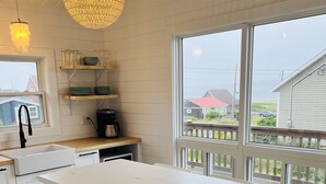 Une cuisine avec une table et des chaises blanches, un luminaire suspendu et une vue sur une maison par une grande fenêtre.