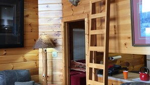 Interior de una cabaña de madera con un ciervo montado en la pared, una escalera de madera, un sofá, una lámpara y un cuadro enmarcado.