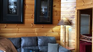 Interior acogedor de una cabaña con una pared de madera, una cabeza de venado montada, cuadros enmarcados, una lámpara y un sofá gris con un cojín.