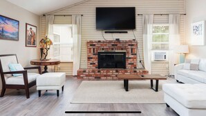 A living room with a brick fireplace, a flat-screen TV mounted above it, a round table with a lamp, and a white sofa.