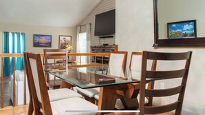A dining area with a glass-top table and wooden chairs, a brick fireplace, a flat-screen TV mounted on the wall, and a framed picture above the fireplace.