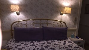 Un letto con copriletto a motivi floreali, due cuscini viola e una testiera dorata. Due lampade a parete su ciascun lato del letto.