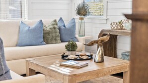 A cozy living room with a beige sofa, blue and green cushions, a wooden coffee table with a book and decorative items, and a rustic wall decor.