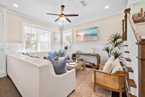 A living room with a beige sofa, wooden coffee table, and a wooden chair. A ceiling fan is visible above.