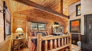Quarto de cabana de toras com uma cama de madeira, uma mesa de cabeceira, um abajur e uma janela com cortinas.