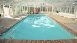 Una piscina interior con paredes de vidrio transparente, iluminada por luz natural, rodeada de sillas de descanso y un expendedor.