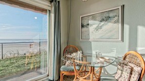 A dining area with a glass table and wicker chairs, a framed artwork on the wall, and a view through a large window.