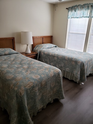 Two single beds with headboards and nightstands, each with a patterned bedspread, are set against a light-colored backdrop.