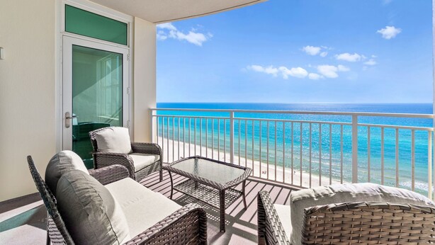 A balcony with wicker furniture and a glass door, overlooking a beach with clear blue water.