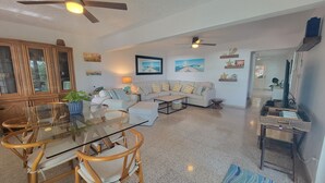 A spacious living room with a glass dining table, wooden chairs, a sofa, and a ceiling fan.