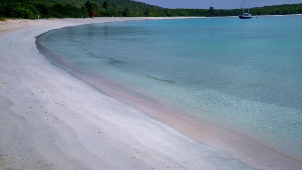 A beach with clear turquoise water, a sandy shore, and a sailboat in the distance.