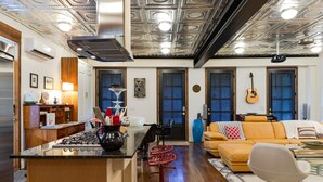 A modern kitchen with a central island, wooden cabinets, and a built-in stove. There is a dining area with a yellow sofa and a white chair. - Porch House W/Swim Spa/Pool &3 Big Screens To Enjoy  (New Orleans)