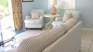A room with a striped sofa, a white armchair, a small round table with a lamp, and a window with blinds. - Direct Oceanfront Cottage-Sunrise View- #2, (Little Torch Key)