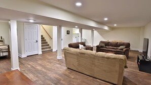 A spacious living room with a large beige sofa, a brown sofa, and a wooden coffee table. A staircase leads to another level, and a television is visible in the corner.