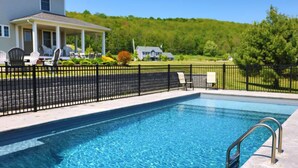 A rectangular swimming pool with a black metal fence, a covered patio area with chairs, and a house in the background.