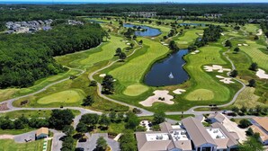 An aerial view of a golf course with a pond, sand bunkers, and a residential area with houses and a small building. - Stunning 6 bedroom 3.5 bath House in Bear Trap Dunes Community! Wifi Available, Pool, Fitness Center, Beach Shuttle, Tennis, Golf! (Ocean View)