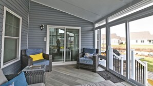 Een overdekt terras met rieten meubels, een schuifpui en uitzicht op een woonwijk.