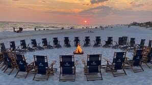 Une plage où des rangées de chaises sont disposées en demi-cercle autour d’un feu central, avec l’océan et le coucher de soleil en arrière-plan.