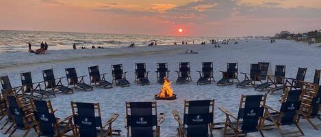 A beach with rows of chairs arranged in a semi-circle around a central fire, with the ocean and sunset in the background.