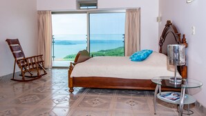 Une chambre avec un grand lit, un fauteuil à bascule, une table en verre et une vue sur la mer.