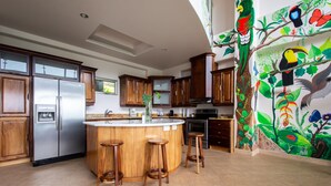 Une cuisine avec des meubles de rangement en bois, un réfrigérateur en acier inoxydable, un îlot central en bois avec des tabourets, et une fresque représentant des oiseaux et des plantes sur le mur.
