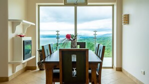 Un coin repas avec une table et des chaises, un vase avec une fleur, et une vue sur un paysage montagneux par le biais d’une grande fenêtre.