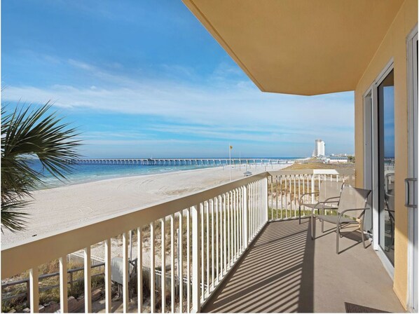 Ein Balkon mit Blick auf einen Strand, einen Steg und ein Gebäude mit Glastür. Eine Palme ist ebenfalls zu sehen.