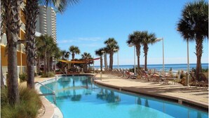 A pool area with lounge chairs and palm trees, surrounded by a building with a balcony overlooking the beach.