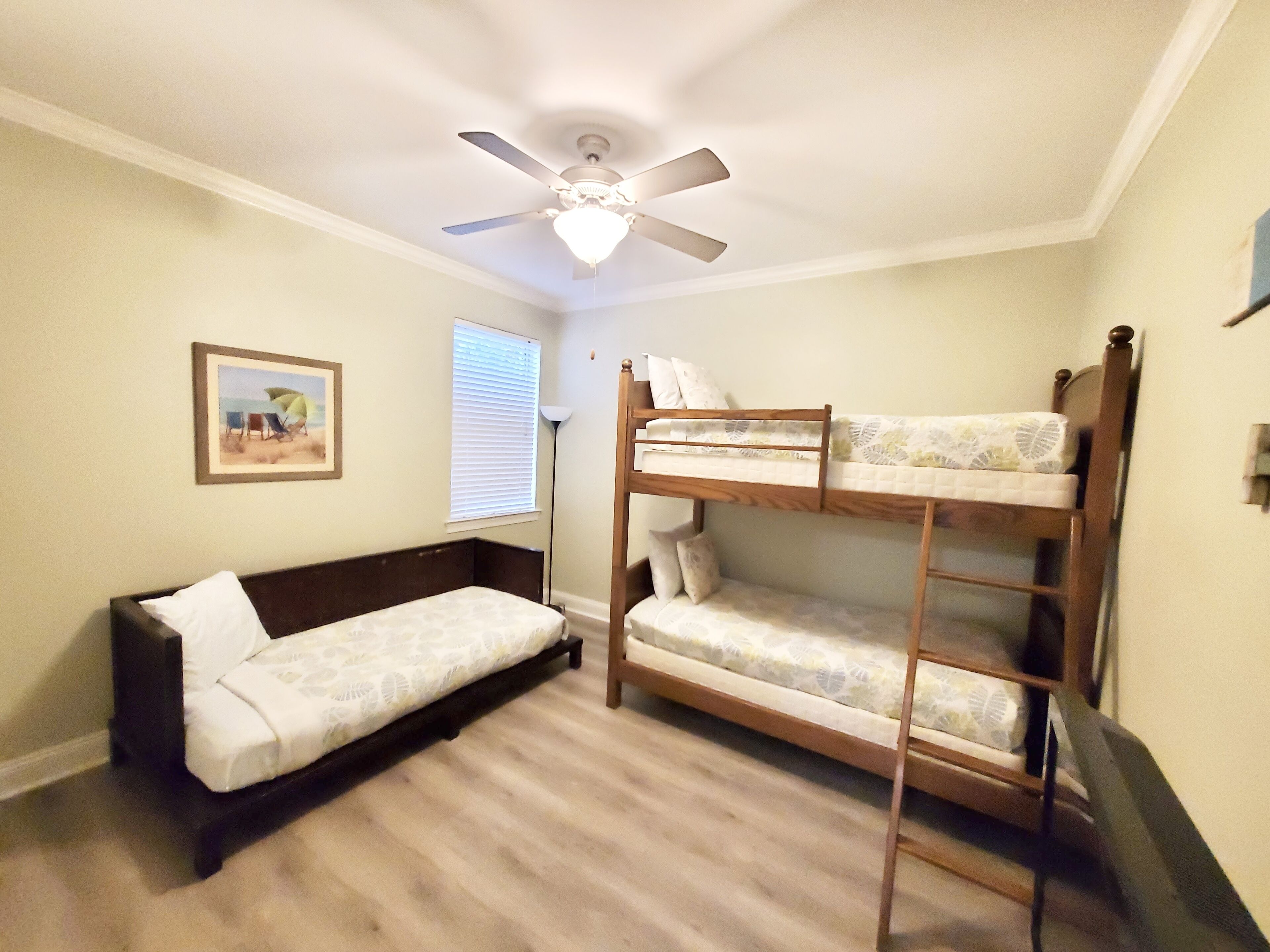 Ein Zimmer mit einem Etagenbett, einer Schlafcouch, einem Deckenventilator, einem Fenster mit Jalousien und einem gerahmten Bild an der Wand.