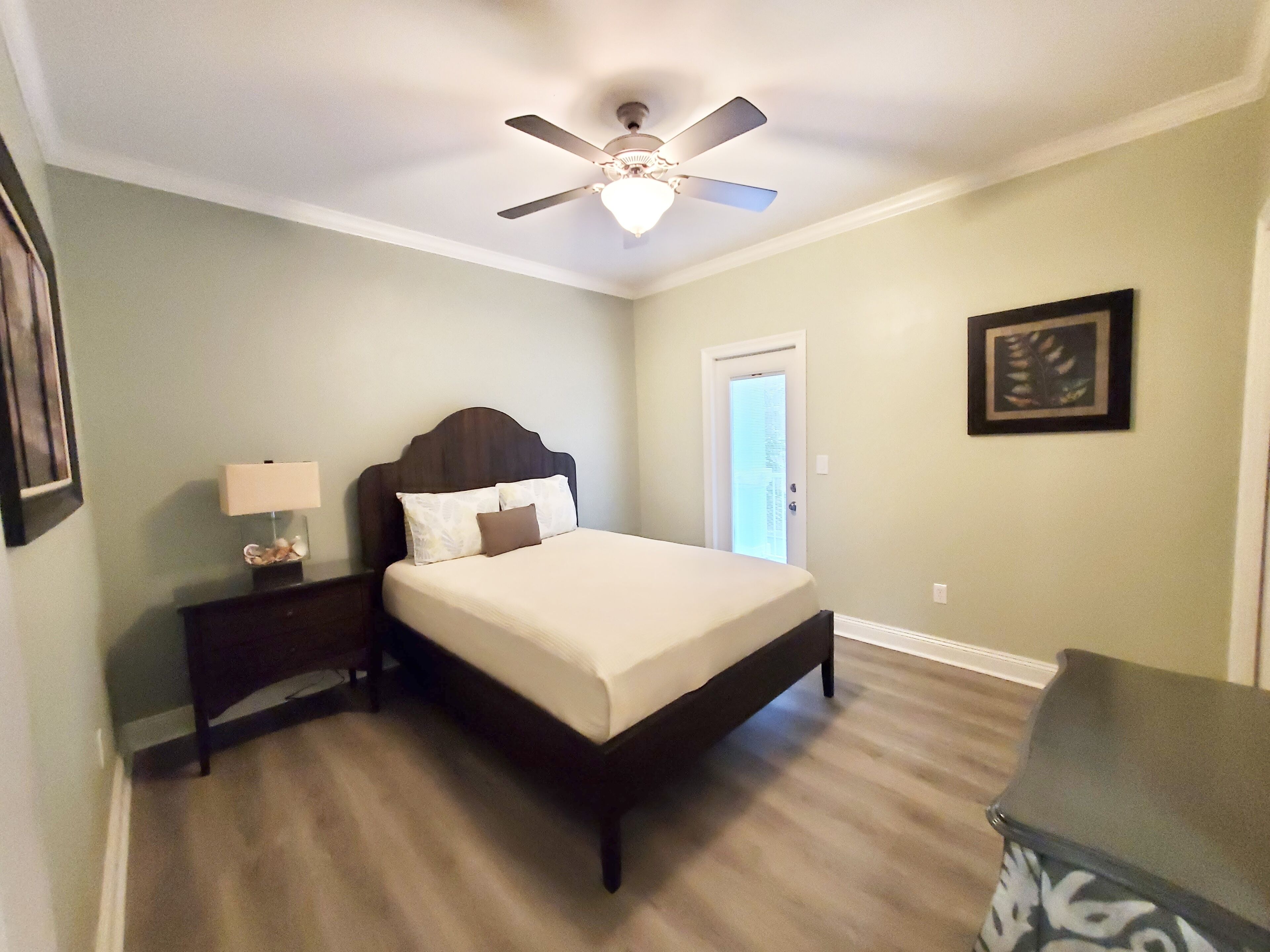 A bedroom with a bed, a nightstand, a ceiling fan, a door, and a framed picture on the wall.