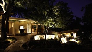 Una casa moderna con un camino de piedra que lleva a una terraza de madera, rodeada de árboles y un brasero de piedra.
