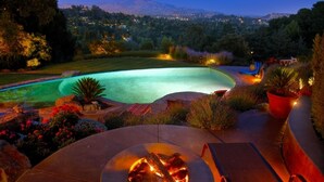 Piscina con un brasero y zona de estar al aire libre, rodeada de jardines ajardinados y vistas a una cadena montañosa.