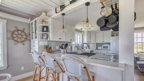 A modern kitchen with white cabinets, a central island with bar stools, hanging pots, and a ship's wheel decoration. - Privacy and Views on Nantucket Island (Nantucket)