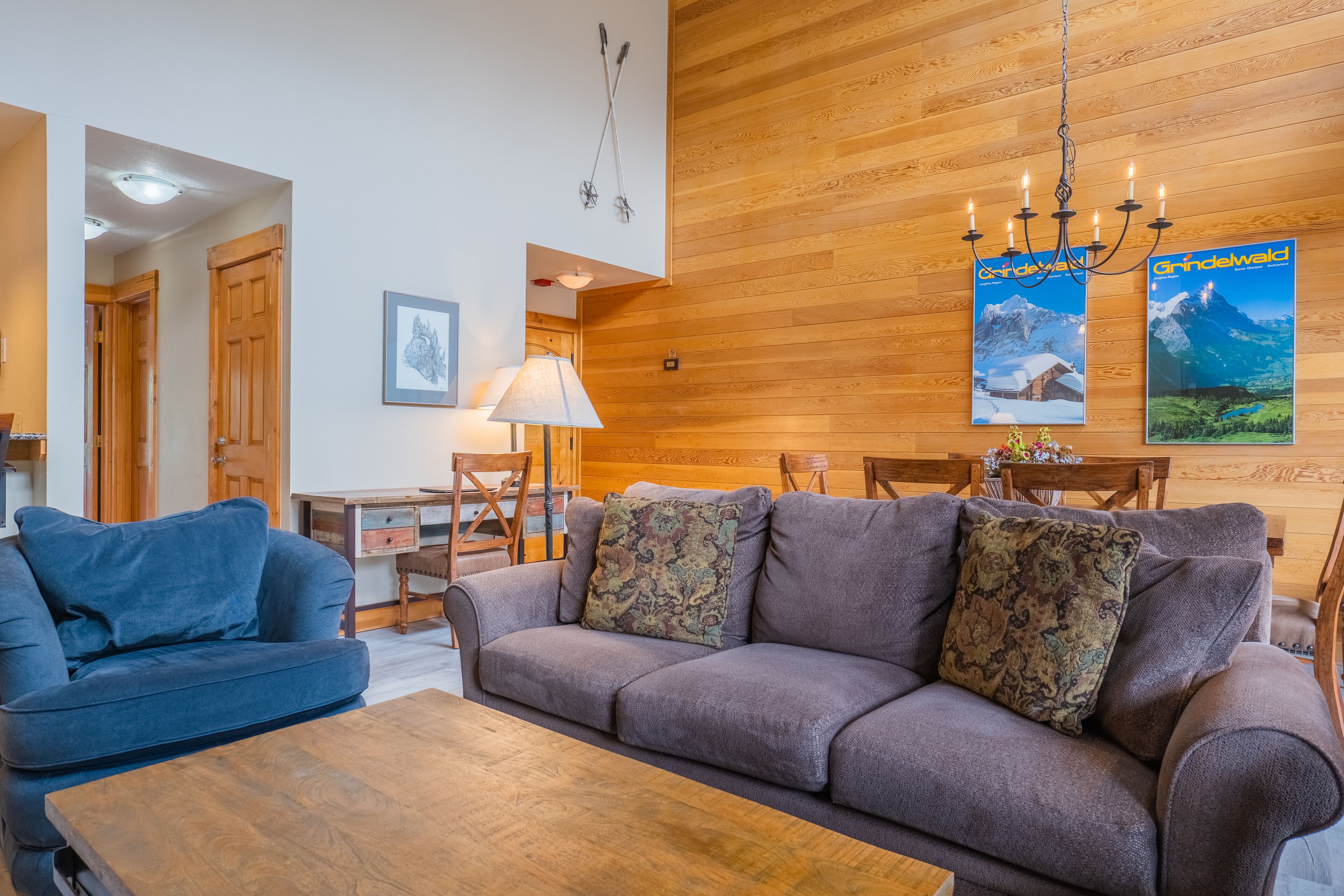 A cozy living room with a sofa, a coffee table, and a wooden wall. There are posters on the wall and a chandelier hanging from the ceiling.