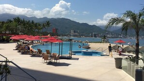A resort pool area with lounge chairs and umbrellas, overlooking a beach and mountains.