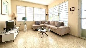 Una sala de estar con un sofá color beige, una mesa de centro de vidrio, un televisor sobre un mueble bajo y una lámpara.