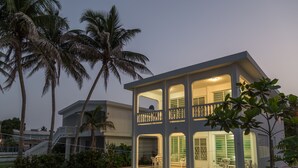 Una casa moderna de dos pisos con balcón, rodeada de palmeras y un césped cuidado.